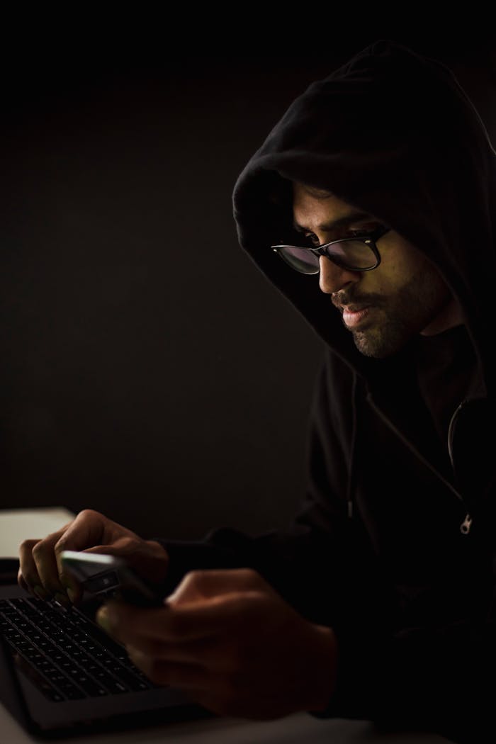 Side view concentrated male hacker in black hood browsing netbook and mobile phone while surfing dark net in obscurity